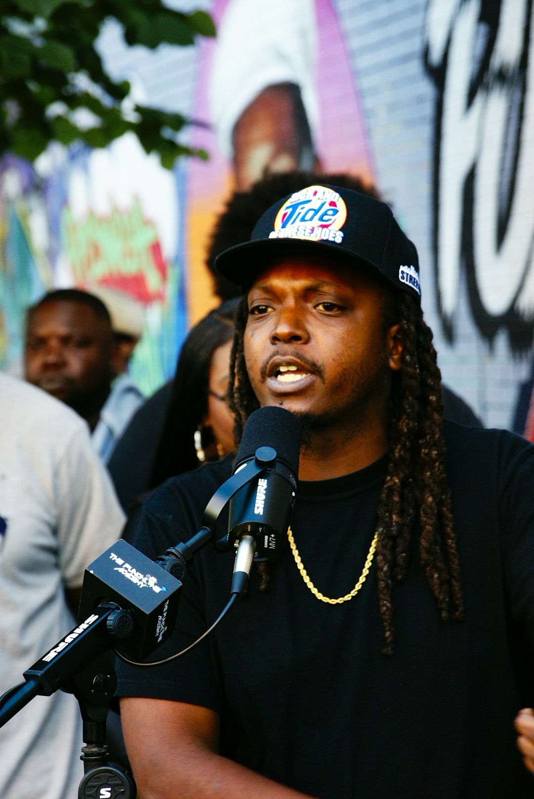 Rah Tha Ruler speaking into a mic in front of a Brooklyn mural